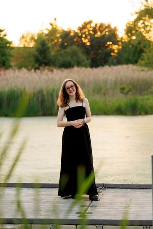 Young woman in black maxi dress smiling on wooden dock at sunset with pond and tall grasses behind her.