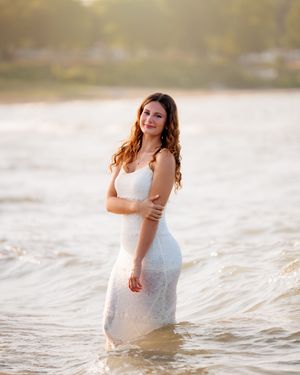 Young woman in white lace dress standing in shallow ocean water at golden hour sunset.