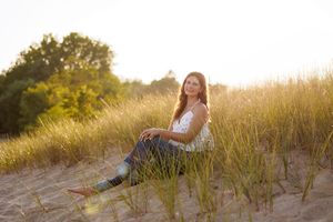 Young woman sitting in golden beach grass dunes at sunset, smiling in white top and jeans.