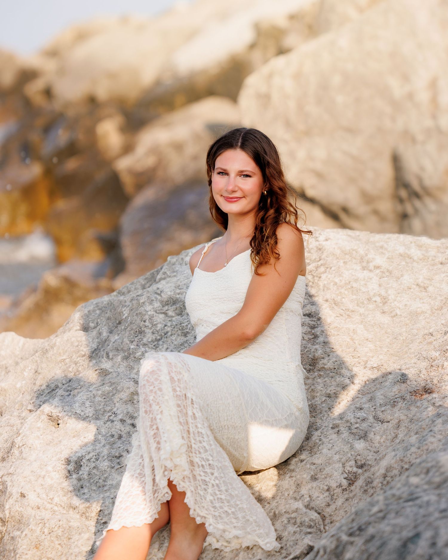 Young woman in white lace dress sitting on rocky beach, smiling warmly with golden sunlight in background.