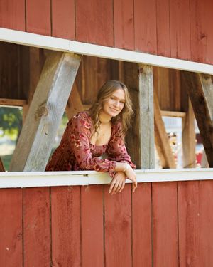 Person in a burgundy top poses creatively through white slats of a rustic red barn wall.