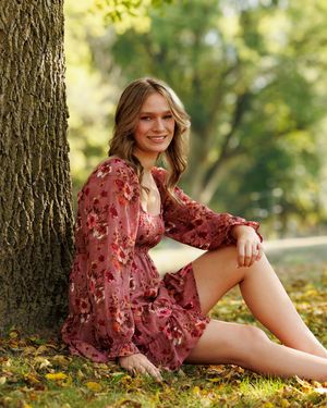 Person in a burgundy floral dress sits against a tree trunk on autumn leaves in soft natural lighting.