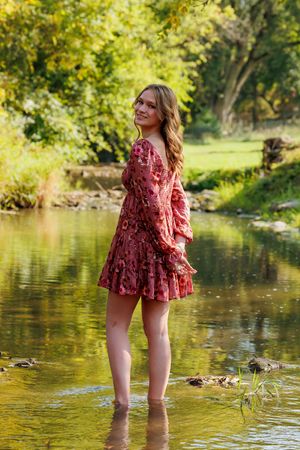 A person in a burgundy floral dress stands barefoot in a shallow stream with sunlit trees in the background.
