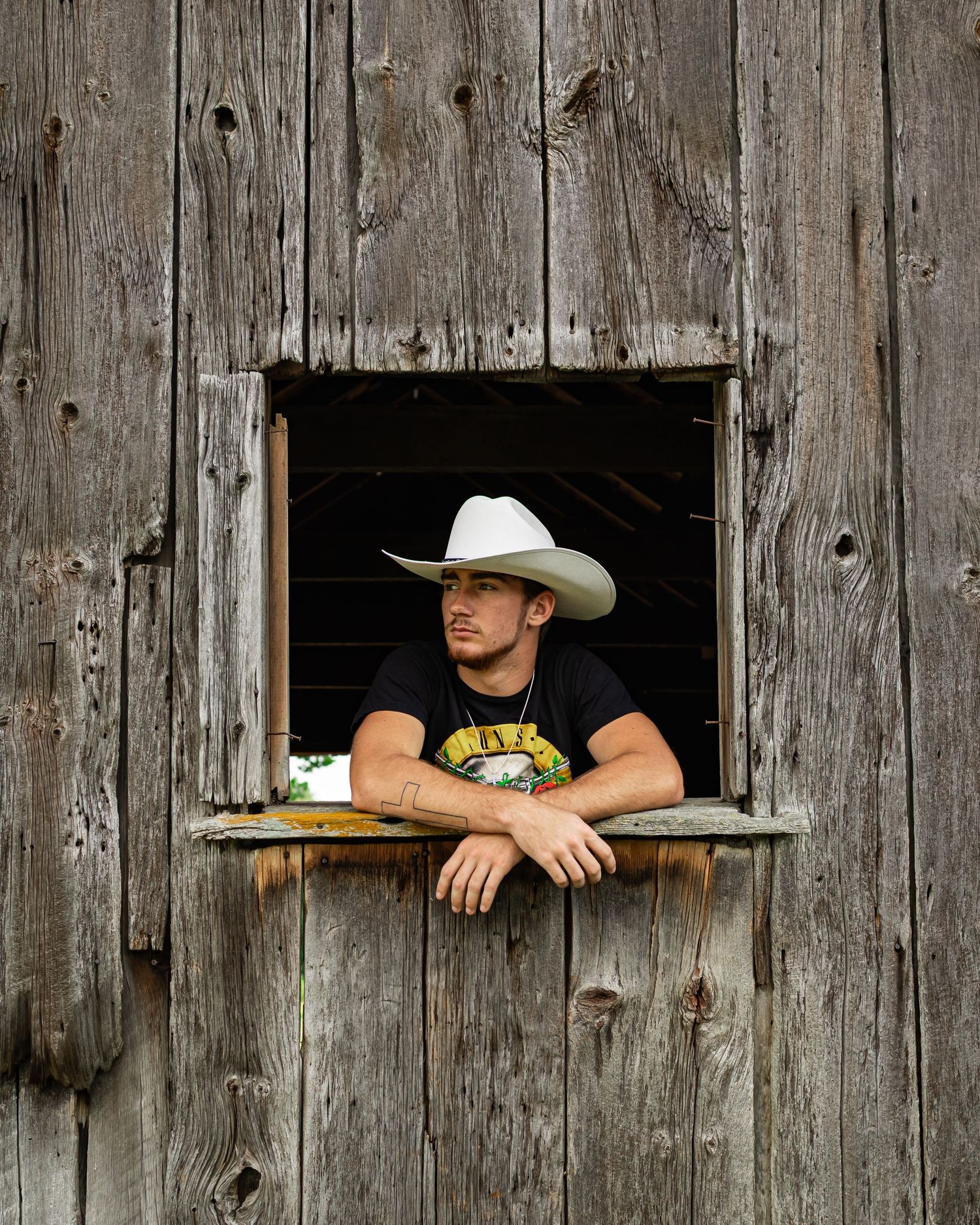 Someone in a black t-shirt and cowboy hat leans through a weathered wooden barn window ledge.