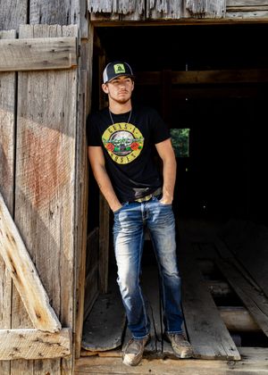 A rustic barn doorway frames someone wearing a Guns N' Roses t-shirt, baseball cap, and distressed jeans.