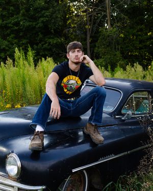 A person in a Guns N' Roses t-shirt sits on the hood of a vintage black car with greenery in the background.