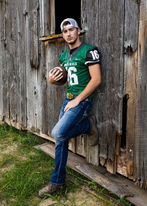A high school football player in a green jersey number 16 poses against a rustic wooden barn door while holding a football.