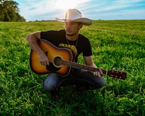 Country musician playing acoustic guitar while sitting in a sunlit green field on a summer day.
