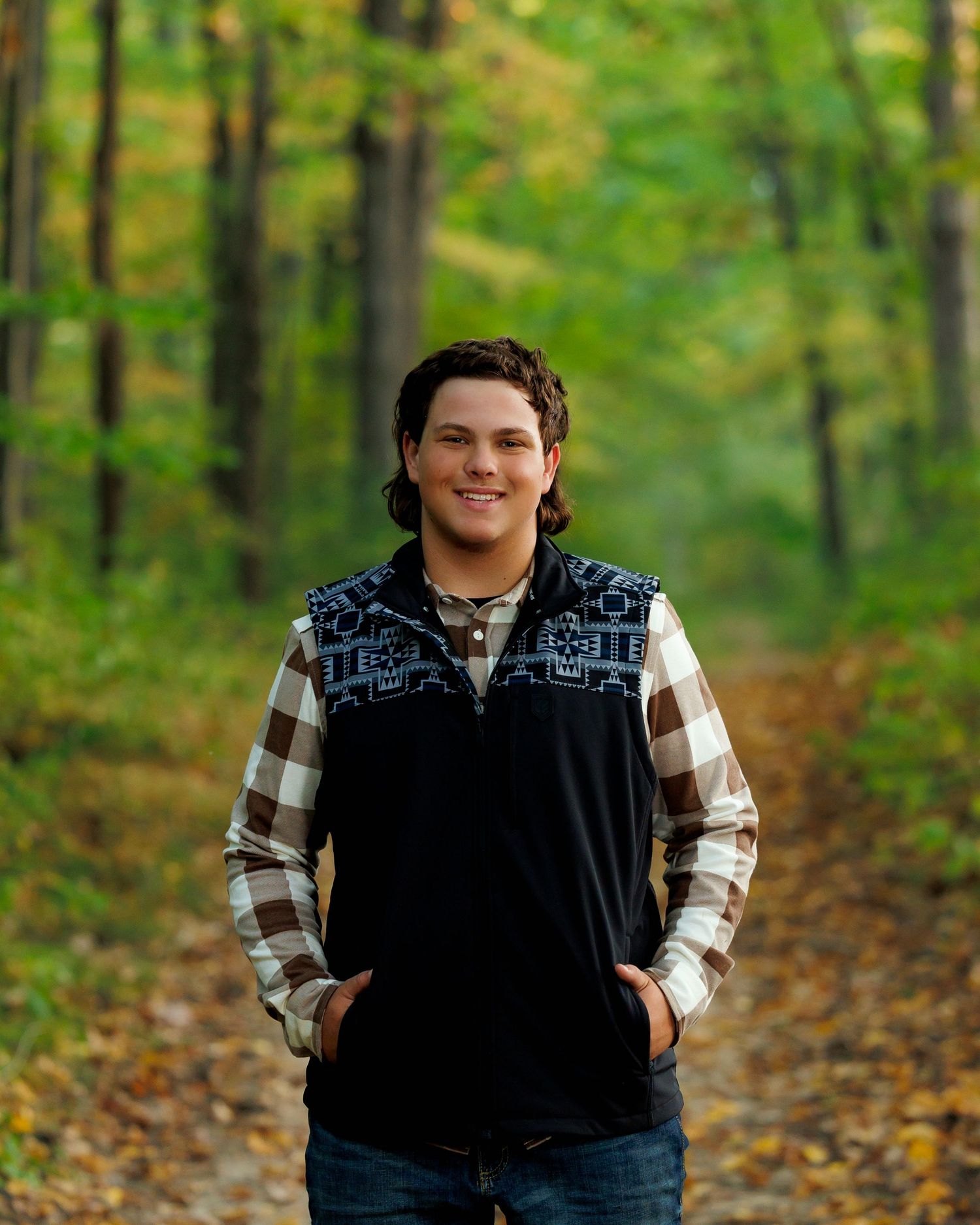 Person wearing plaid shirt and black vest poses in autumn forest setting with fallen leaves on ground.