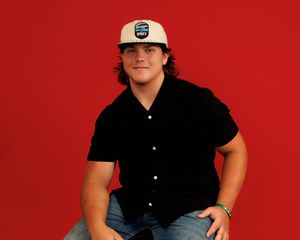 Person wearing a black shirt and baseball cap poses against a red background in a casual seated position.