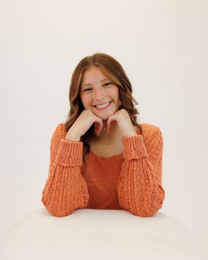 Smiling highschool senior in a cozy orange cable knit sweater rests their chin on their hands against a white background.