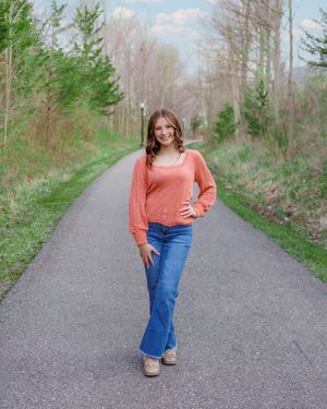 A high school senior poses casually on a paved trail surrounded by spring trees while wearing a coral sweater and boot-cut jeans.
