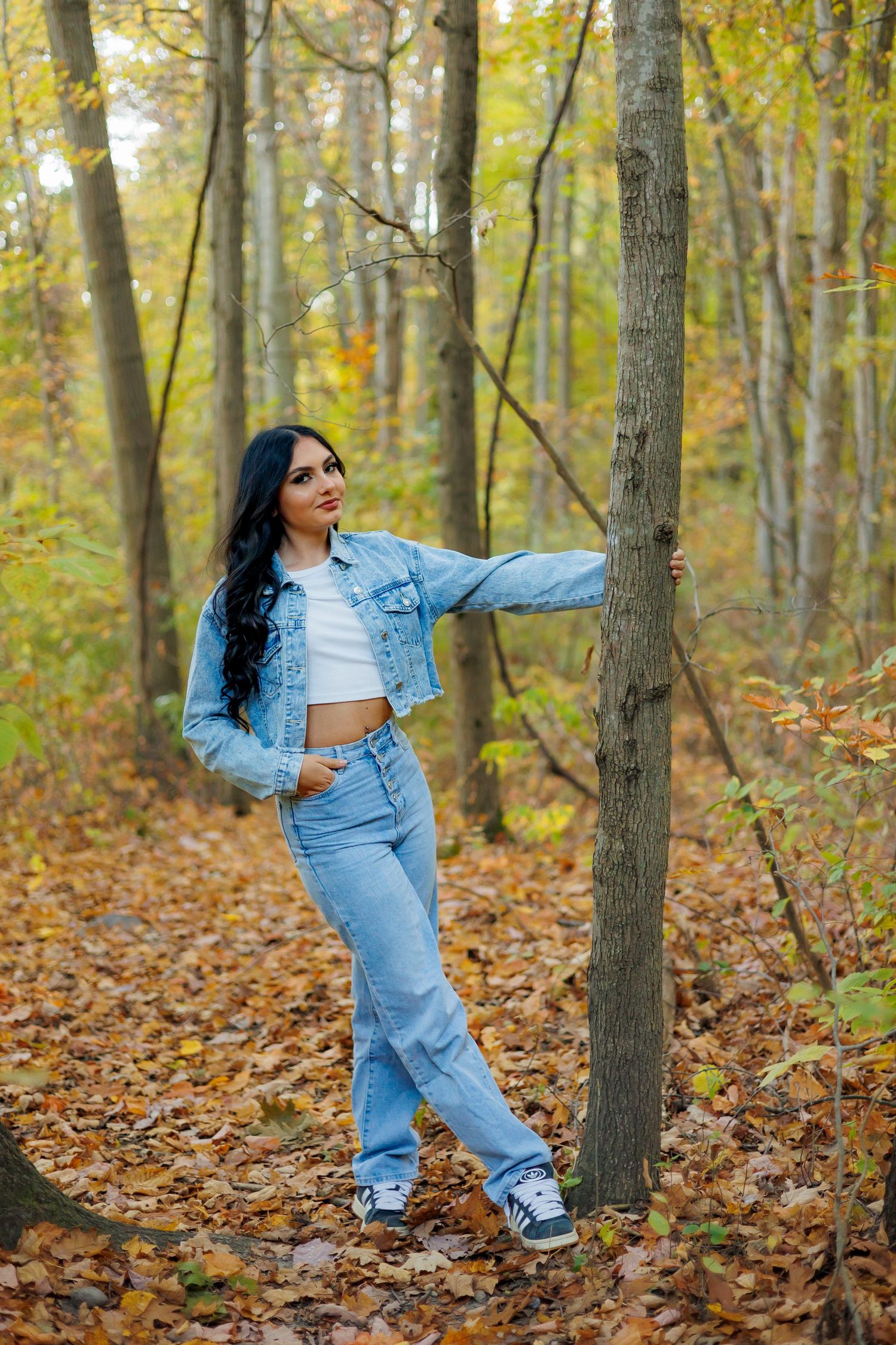 A figure in denim poses gracefully against autumn trees with fallen leaves scattered on the forest floor.