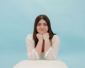 A person in a white lace dress rests their chin on their hands against a light blue background.