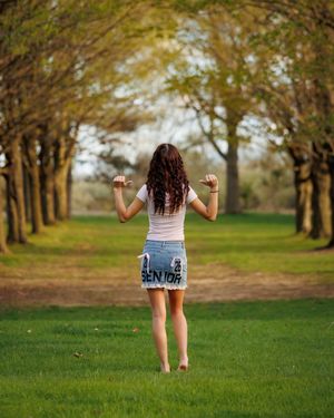 A girl with curly hair wearing a denim skirt stands barefoot on a grass path lined with tall trees.