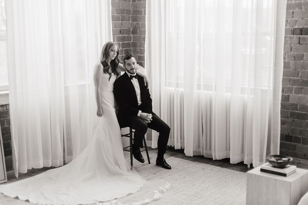 A black and white wedding photo of a couple posing in front of sheer curtains and brick walls.
