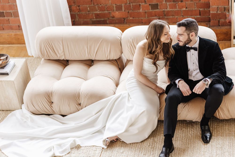 Couple in formal wedding attire sitting together on a beige couch against exposed brick wall.