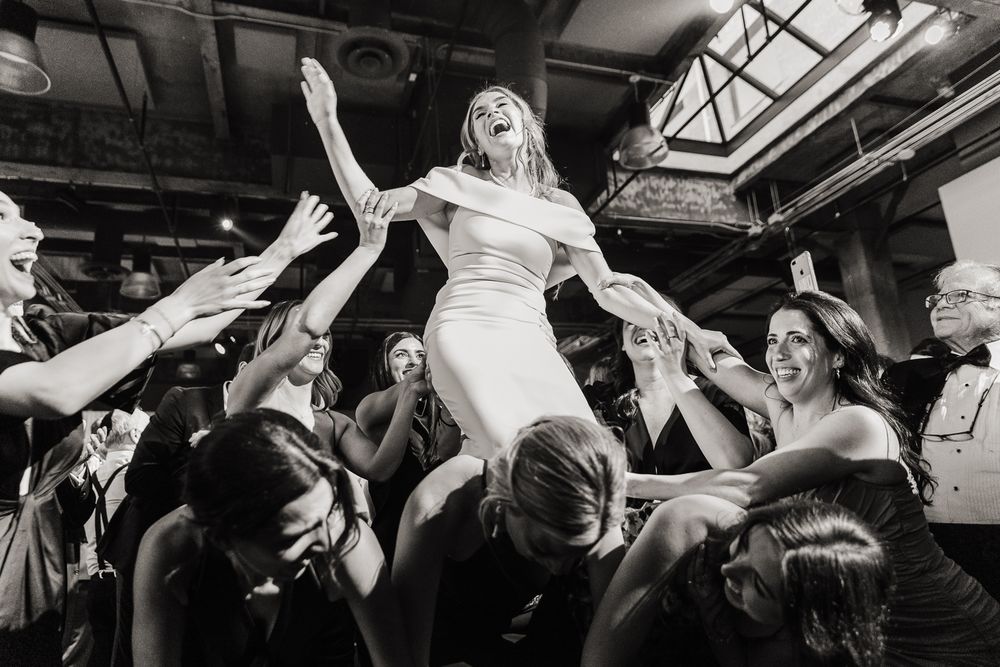 A bride in a white dress is lifted and celebrated by wedding guests during a joyous reception dance moment.