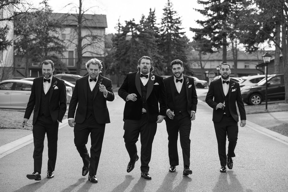 A group of groomsmen in black suits walk together on a street in black and white photography.