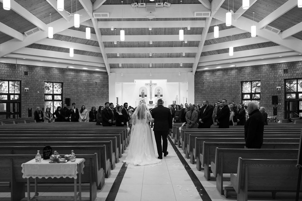 Black and white view down the center aisle of a church during a wedding ceremony with guests seated in wooden pews.
