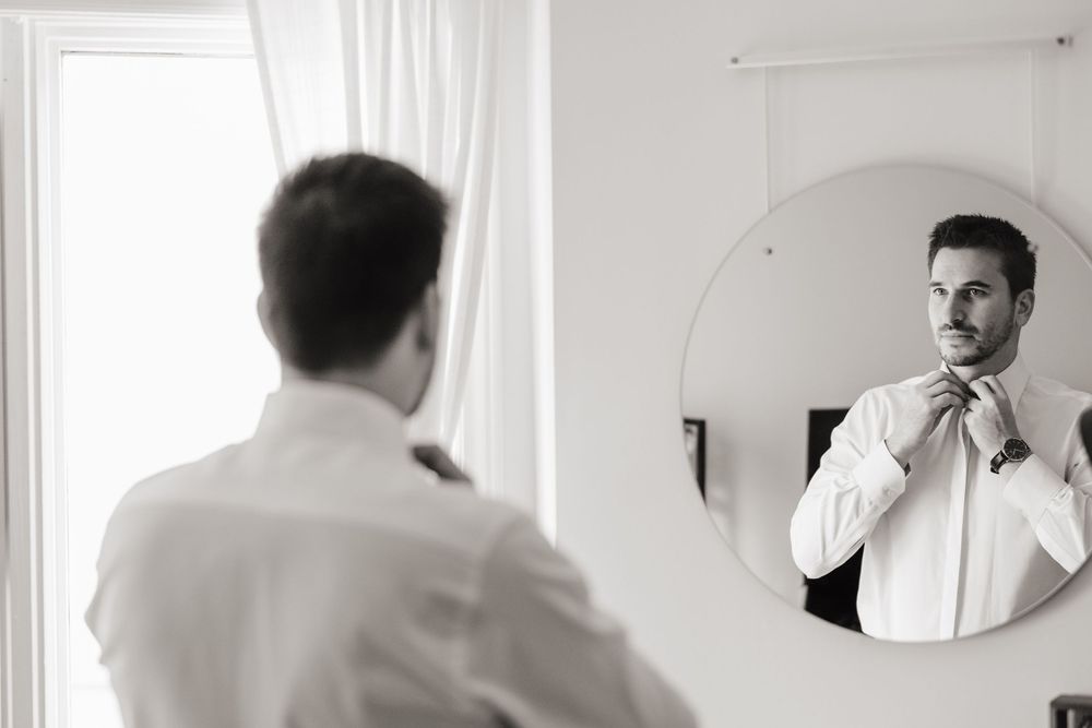 A person adjusts their white shirt collar while looking into a circular mirror in a bright room.