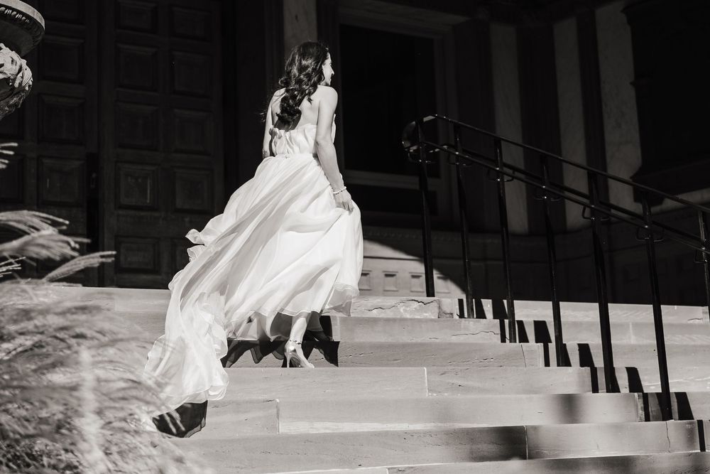 A bride in a flowing white wedding dress descends a grand staircase in dramatic black and white photography.