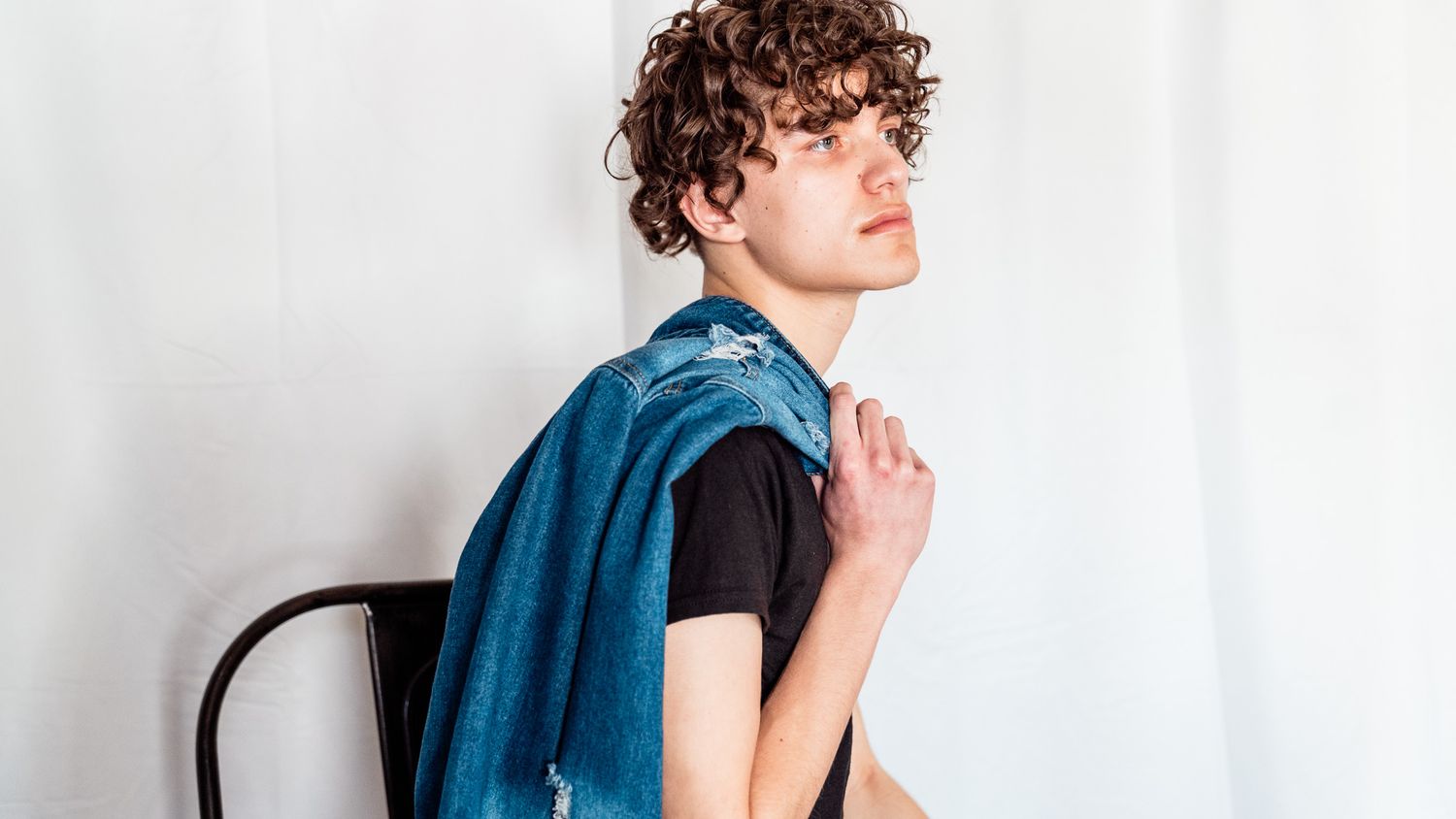Young person in black t-shirt with curly hair sits casually against chair with blue fabric.