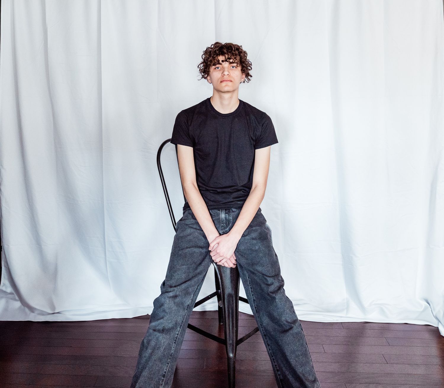 Person in dark t-shirt and cargo pants poses on chair against white backdrop.