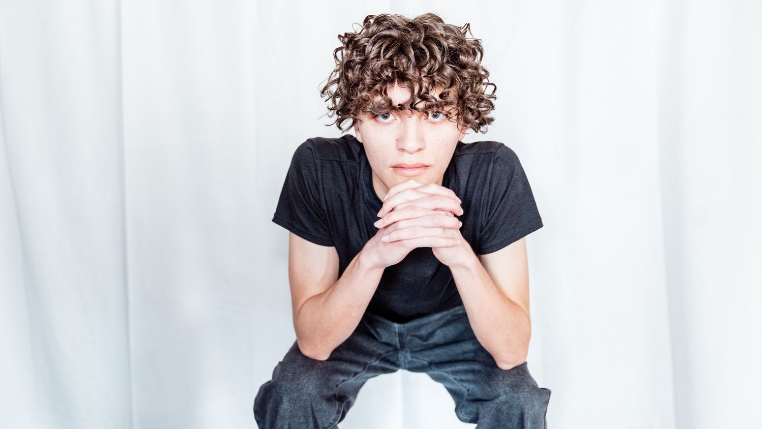 Person in black t-shirt and gray pants rests head on clasped hands while seated.