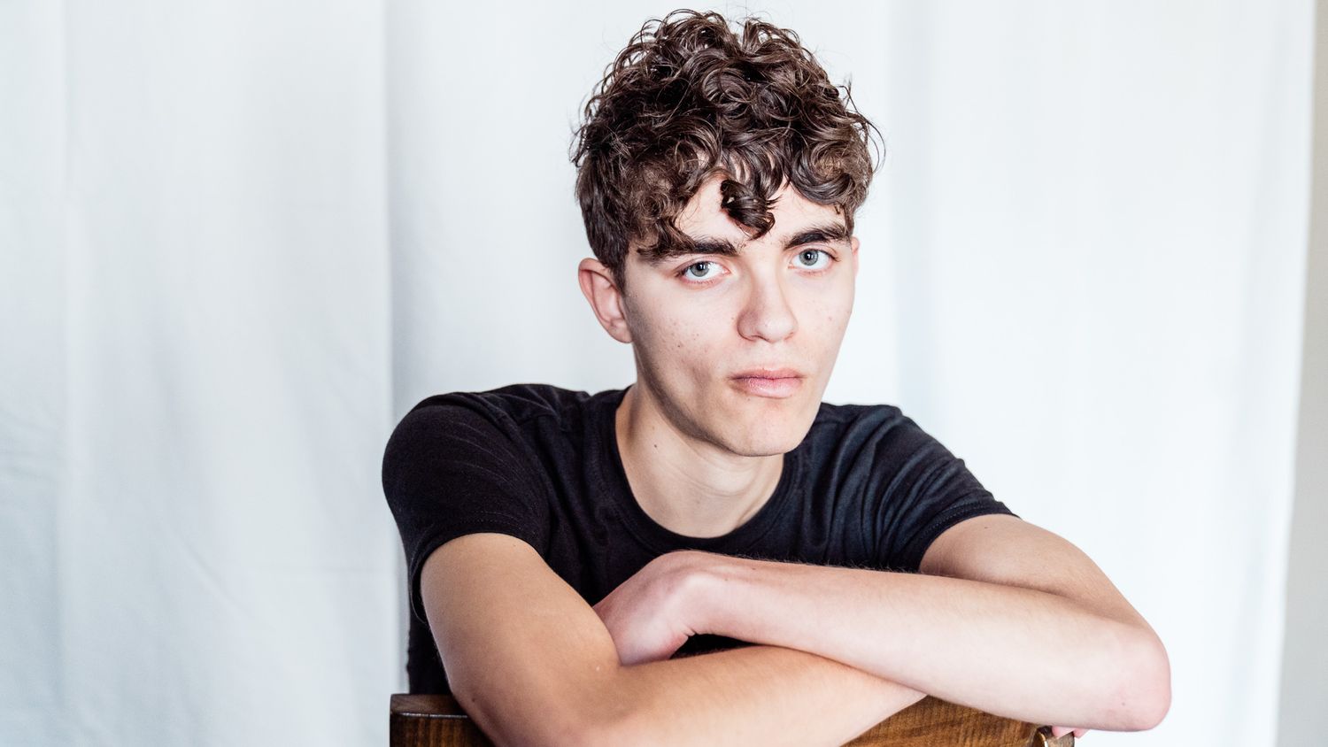 Portrait of person in black t-shirt leaning on wooden surface against white background.