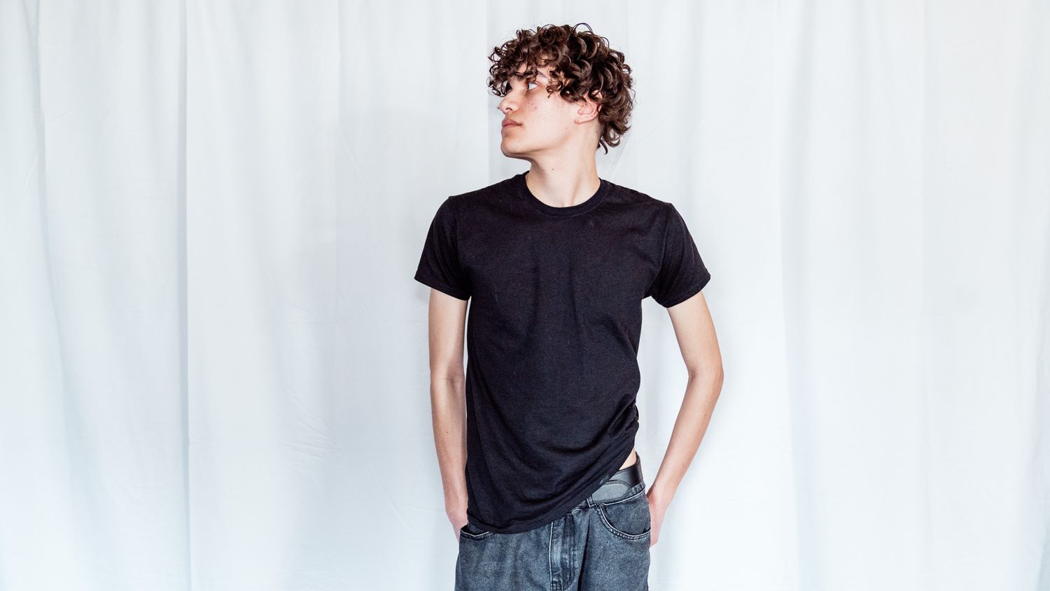 Person in black t-shirt and gray pants stands casually against white backdrop.
