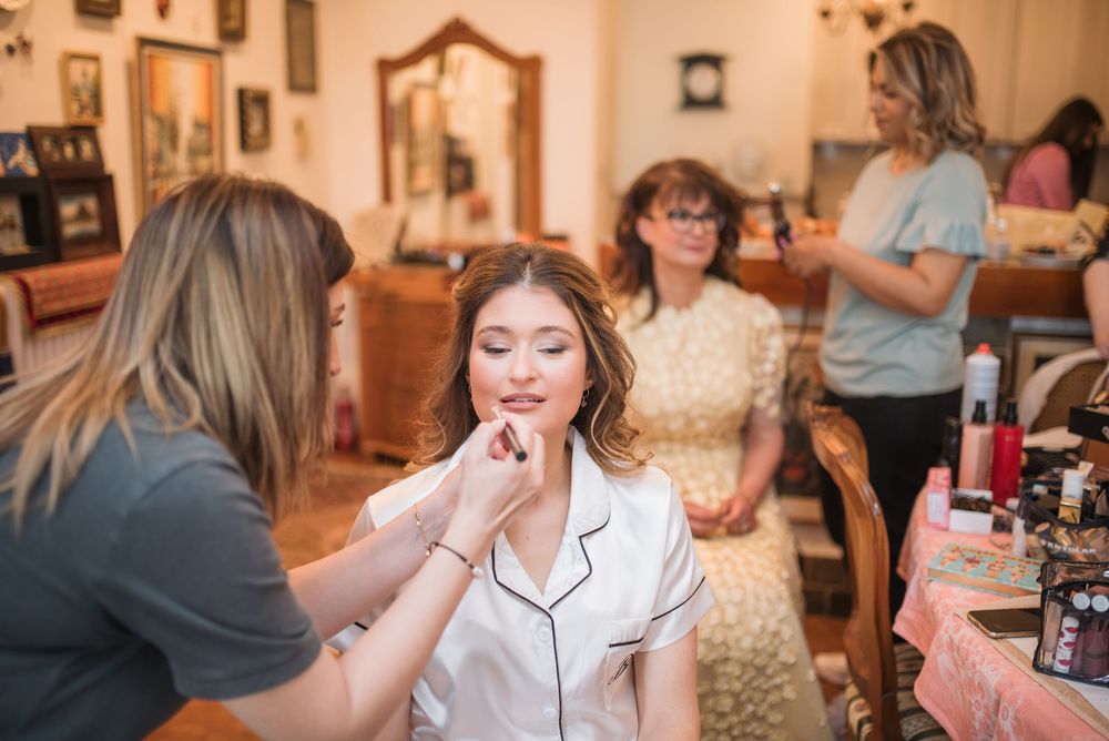 Professional makeup artist applying cosmetics to bride wearing white silk robe during wedding day preparation.