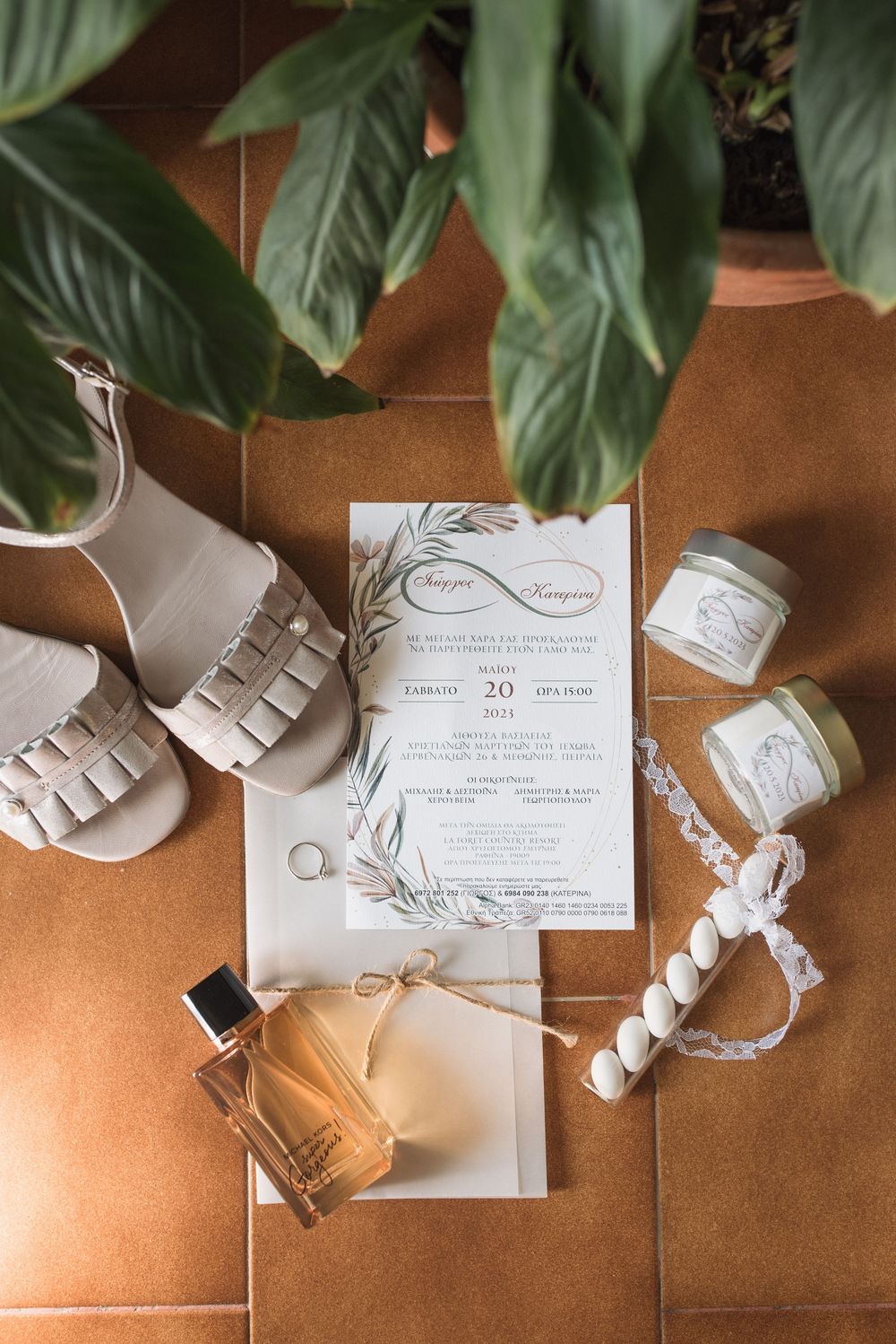 Wedding details flatlay with invitation, shoes, perfume and green leaves on terracotta background.