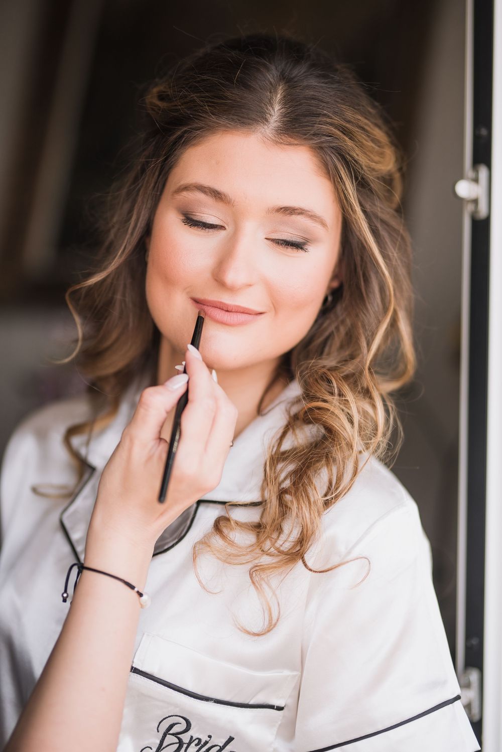 Bride in white monogrammed pajamas having lip makeup applied during beauty preparations.