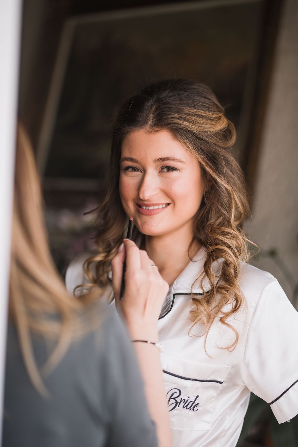 Bride in white monogrammed pajamas getting makeup applied during wedding preparations.
