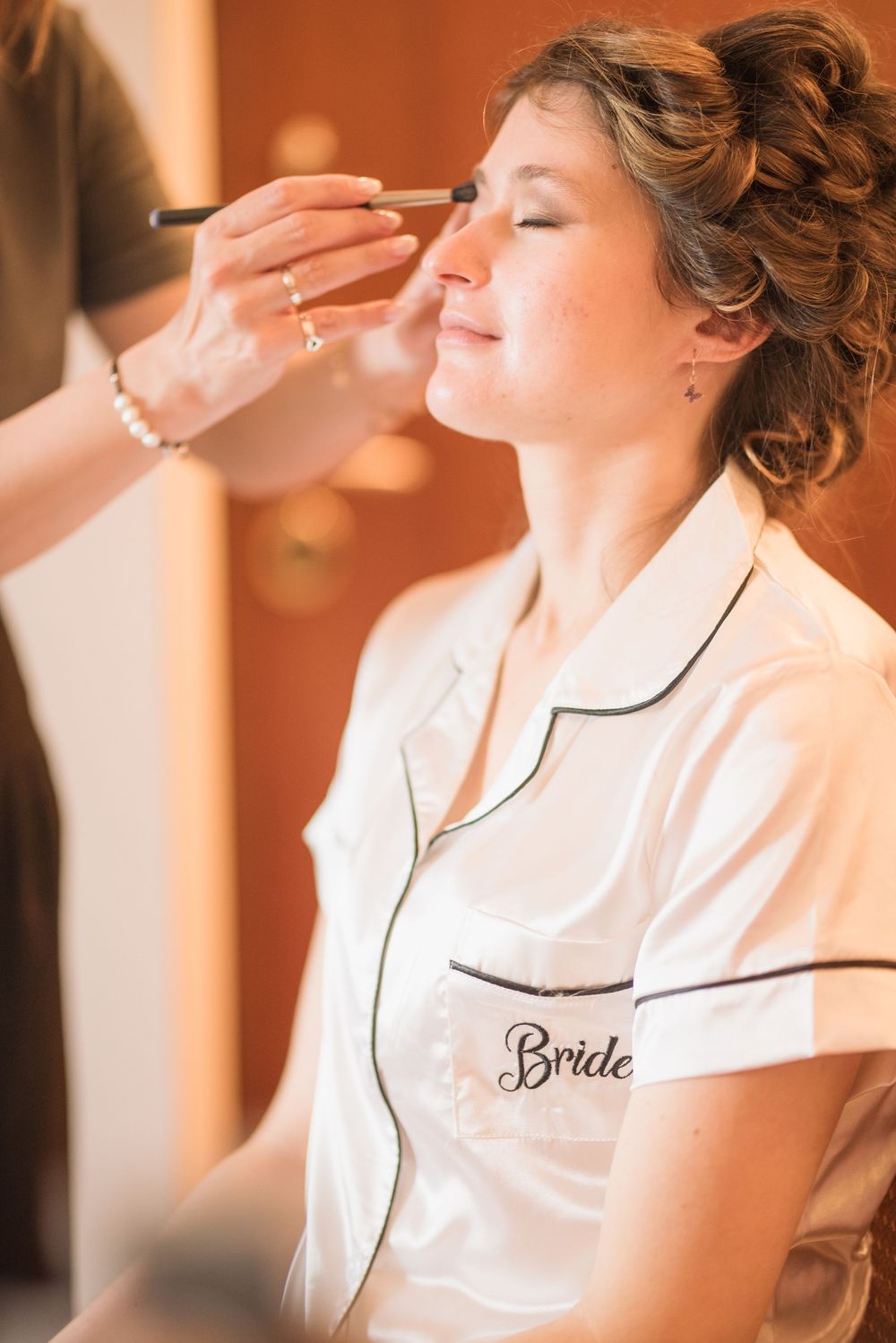 Makeup artist applying cosmetics to bride wearing white silk robe during wedding preparations.