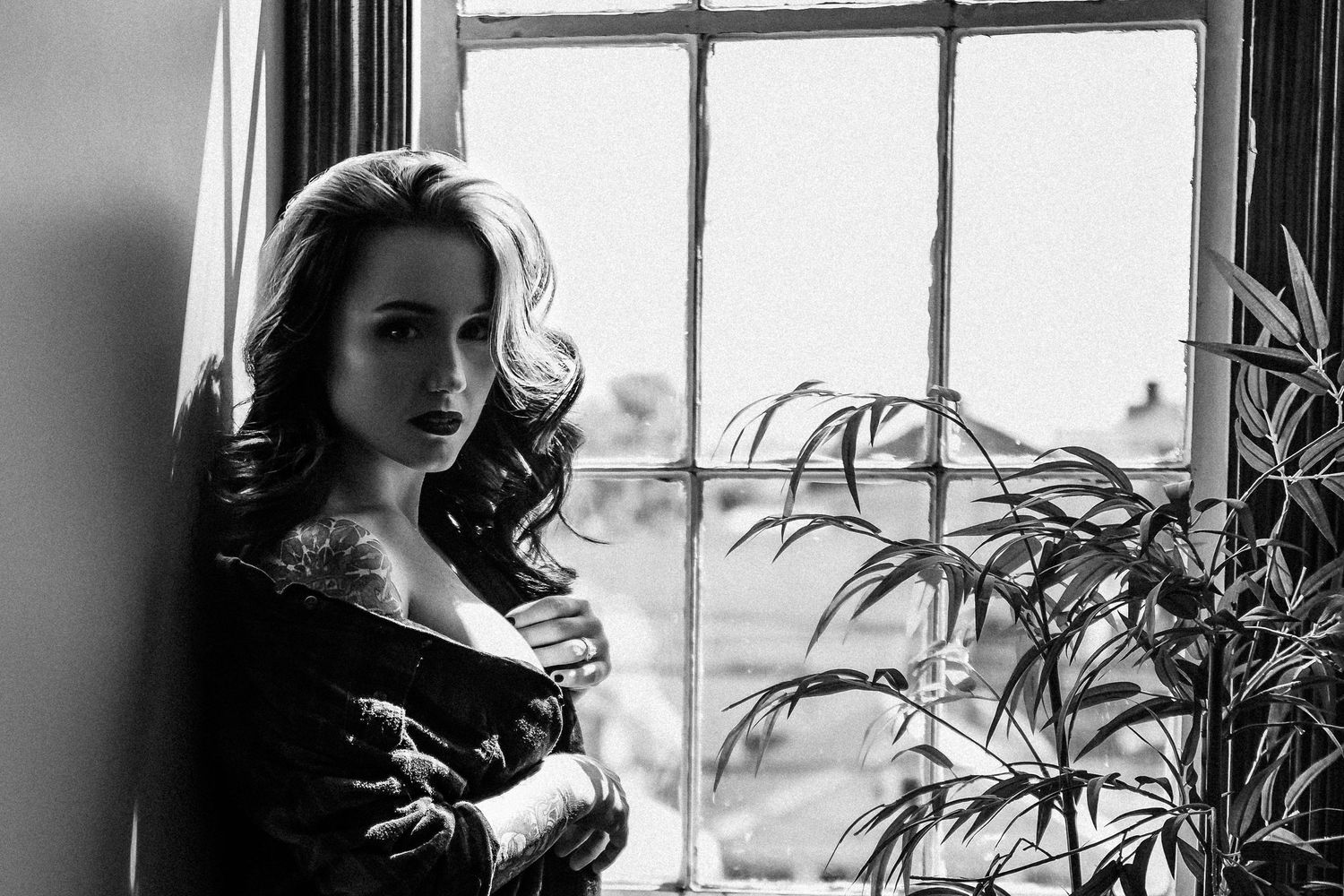 Black and white portrait of a woman with curly hair posing near a window with a plant beside her. soft boudoir photoshoot
