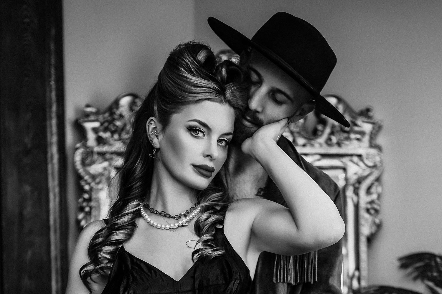 Black and white portrait of an elegant woman in pearls and a man in a hat posing together in a vintage setting. soft boudoir photoshoot