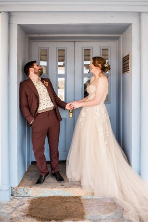 Wedding couple sharing a joyful moment in front of a blue doorway while dressed in burgundy suit and flowing white gown.