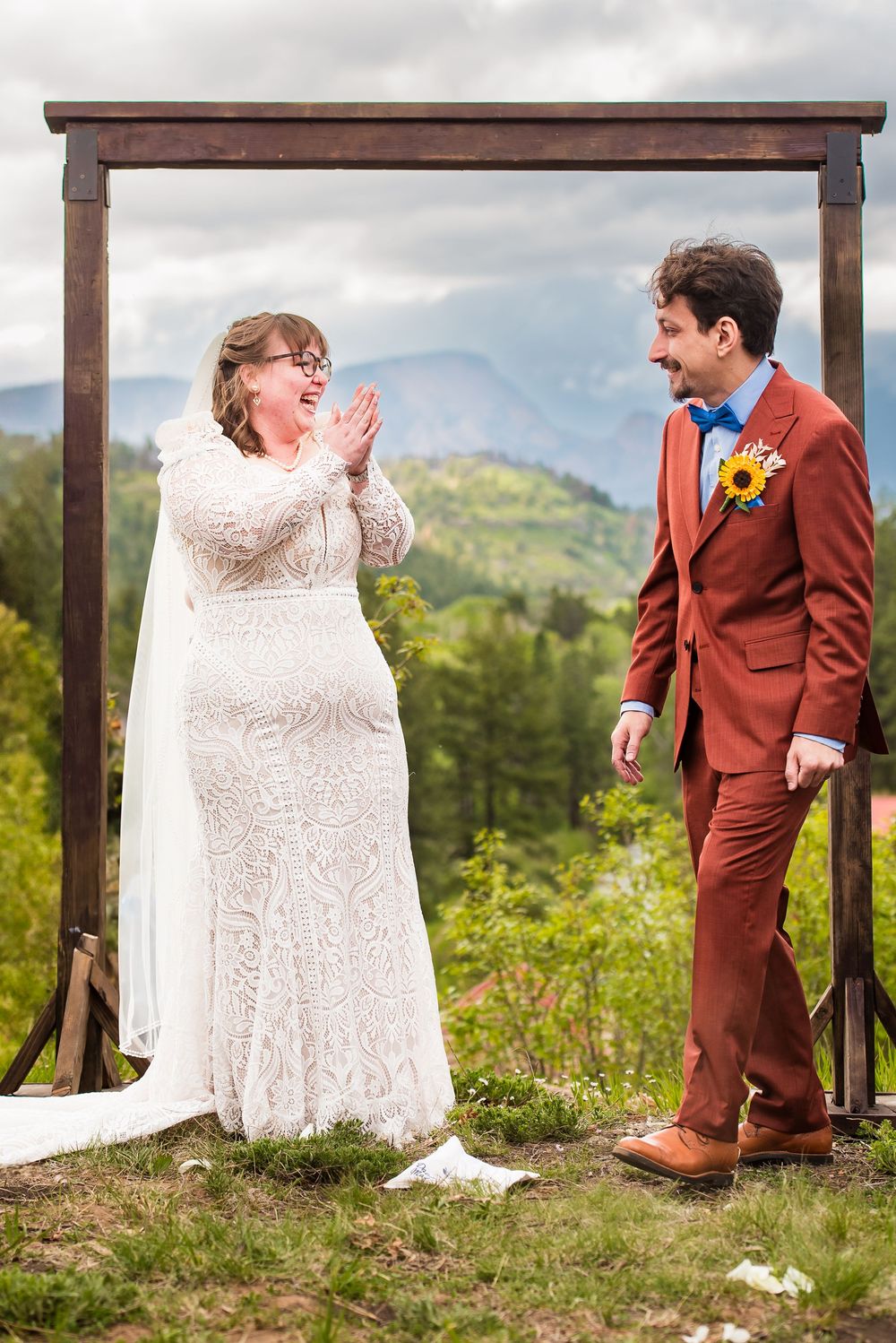 A bride in a lace gown and groom in rust-colored suit share a joyful moment under a wooden arch in the mountains.