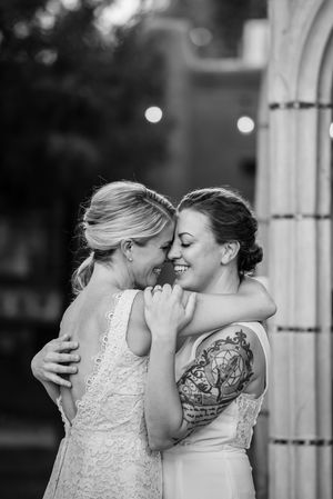 Two brides share an intimate moment in white wedding dresses, one with a floral arm tattoo, during their outdoor ceremony.