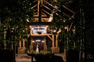 A couple shares a romantic moment under an illuminated rustic wooden entryway at night surrounded by trees at Blame Her Ranch in New Mexico.