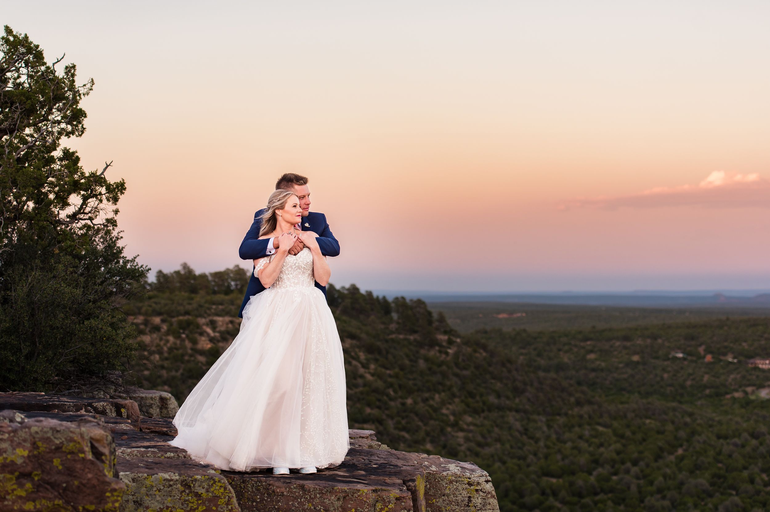 A couple in wedding attire embraces on a rocky overlook during sunset.