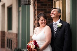 Joyful couple laughs together against brick building while bride holds red bouquet at Hotel Andaluz in Albuquerque, New Mexico.