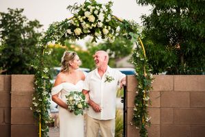 A romantic wedding ceremony under a floral arch adorned with white and yellow blooms in an outdoor garden setting.