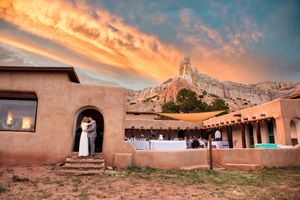 A rustic adobe with string lights at sunset beneath dramatic orange clouds and mountain peaks in the background at the Historic Ghost Ranch.