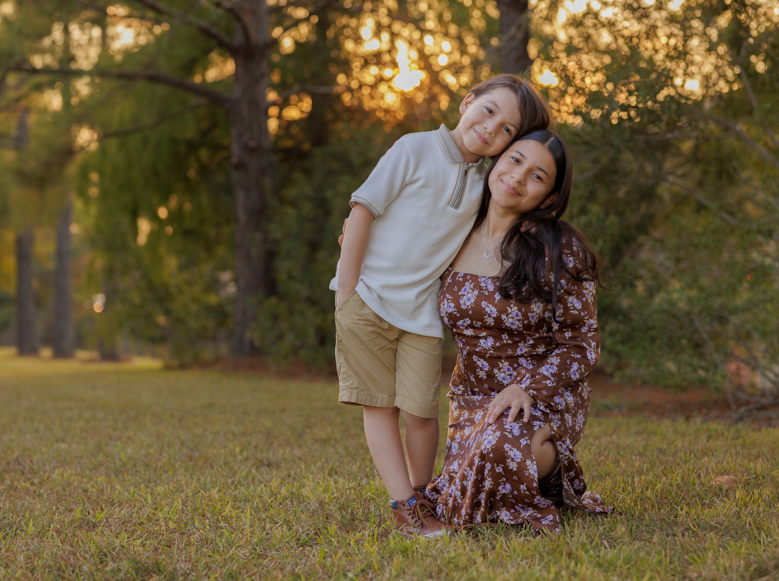 A couple sits together on grass in a park during golden hour sunset with trees in the background.