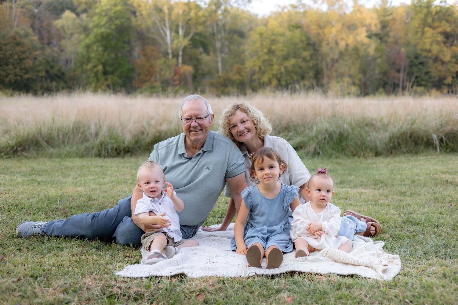 Family lying together on white blanket in grassy field during autumn photoshoot.