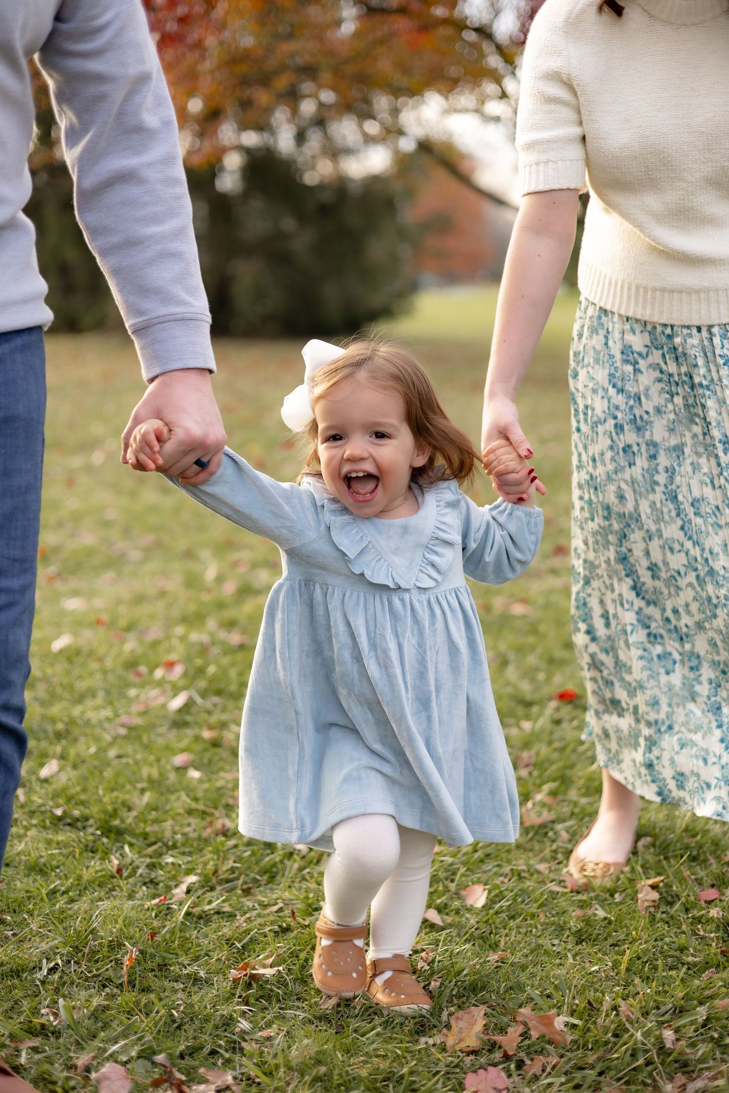 A child in a light blue dress walks through autumn grass while holding hands with adults in a candid moment.