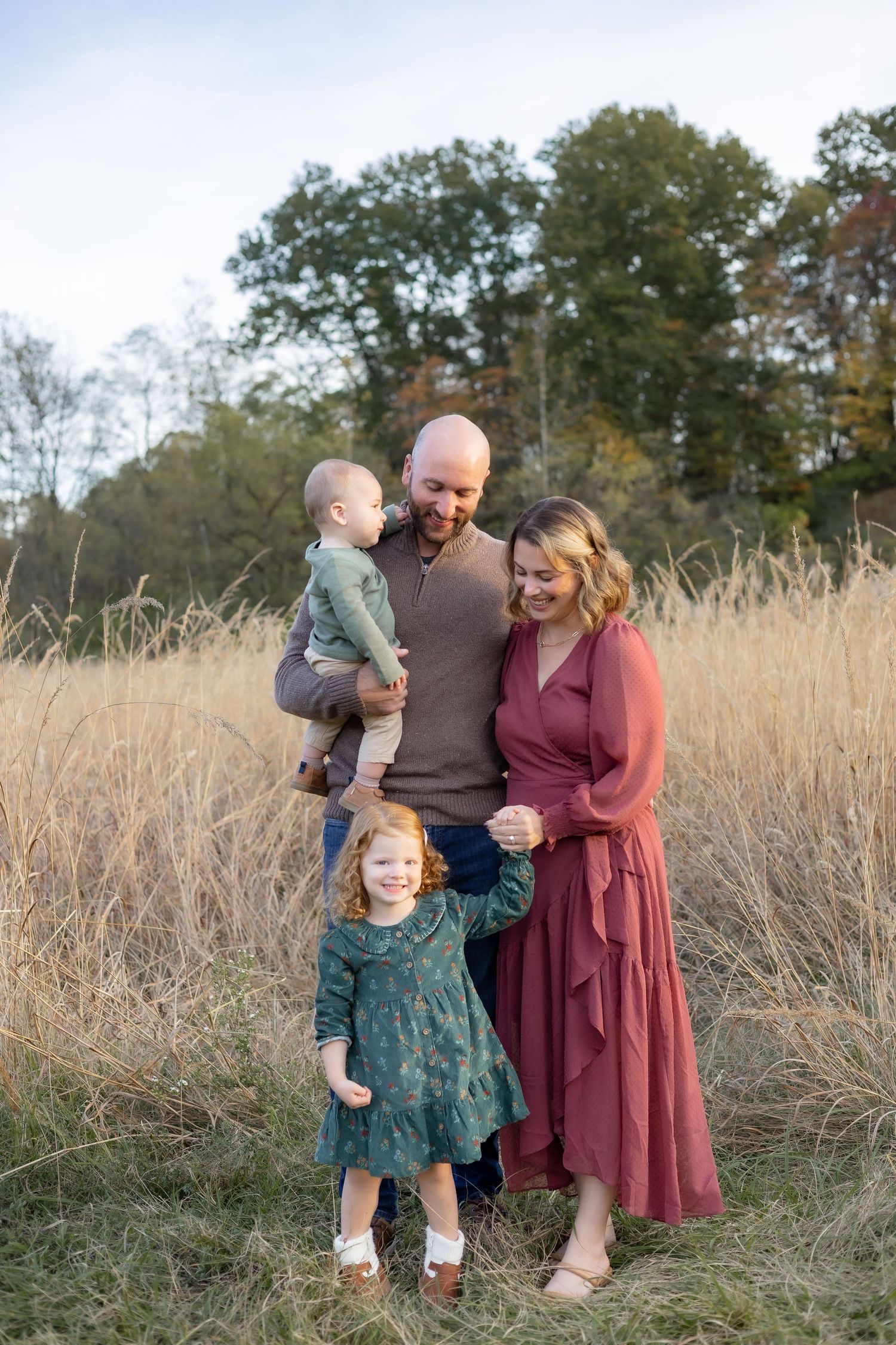 A family stands together in a golden autumn field with tall grass and trees in the background.